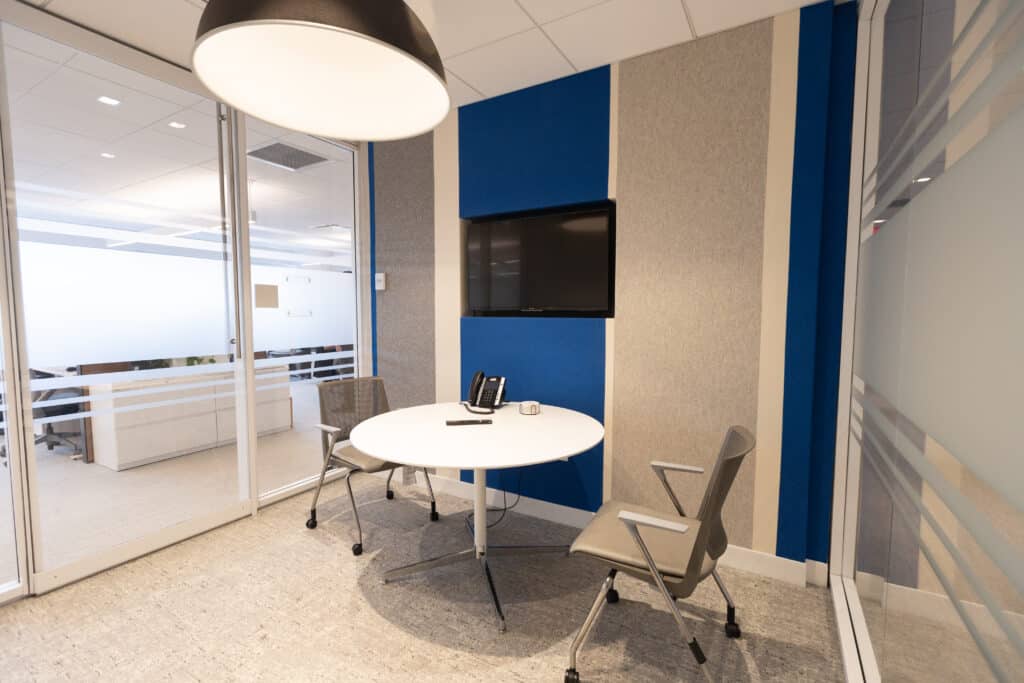 Small glass-walled meeting room at LGRA with round table, two chairs, wall-mounted screen, and blue accent wall